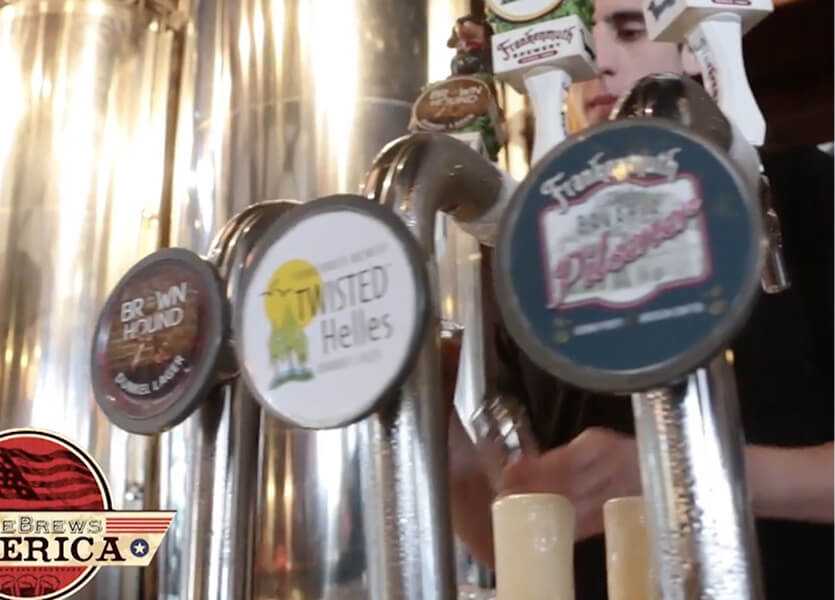Close-up of beer taps at a bar with different labels, including Brown Hound, Twisted Helles, and Funktober Fest Lager. A bartender is blurred in the background filling glasses.