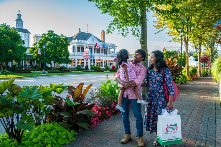 A family stands on a flower-lined sidewalk near a historic white building with flags. The father holds a young child, while the mother carries a shopping bag. Trees and colorful plants surround them on a sunny day.