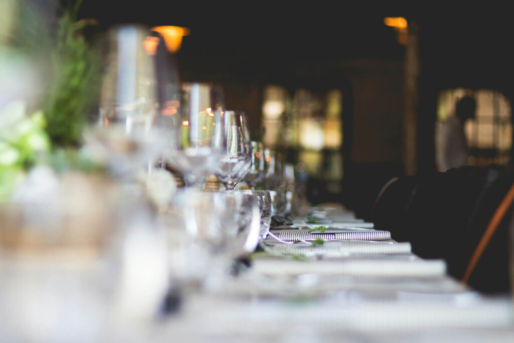 A close-up view of an elegant dining table set with wine glasses, plates, and napkins, softly lit in a dimly lit restaurant or event setting. The background is blurred, emphasizing the table arrangement.
