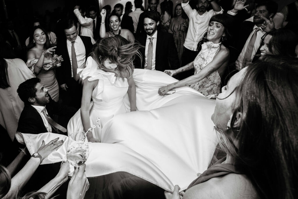 A joyful bride in a white dress dances surrounded by friends and guests, who are lifting her dress and celebrating together at a lively wedding reception.