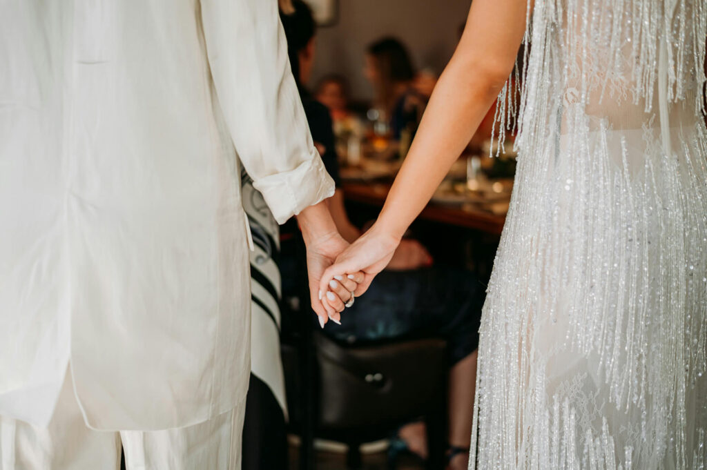 Two people, one in a white suit and the other in a sparkly dress, are holding hands and standing side by side, seen from behind, with a blurred background of people seated at a table.
