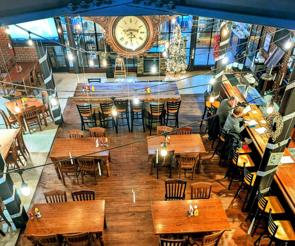 A warmly lit restaurant with wooden tables and chairs, viewed from above. A large vintage clock and decorated Christmas tree are against the back wall. A few people sit at separate tables and at the bar. Hanging lights add a cozy ambiance.