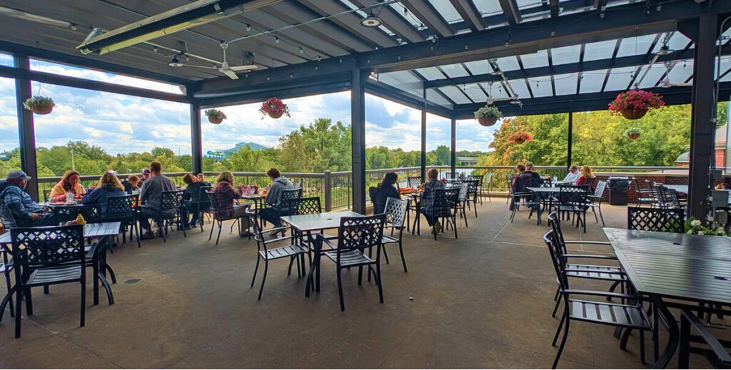 People dining and chatting on a spacious, covered outdoor patio with black metal tables and chairs, surrounded by hanging flower baskets and overlooking trees under a partly cloudy sky.