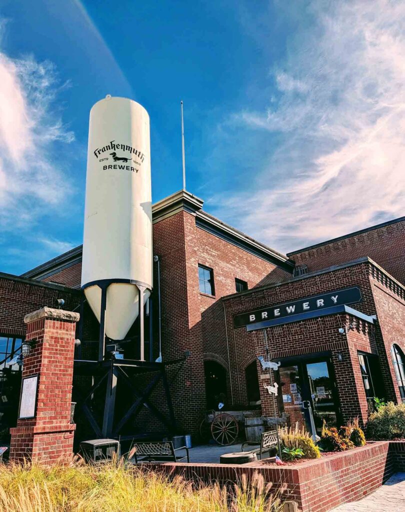 Exterior view of the red-brick Frankenmuth Brewery building with a large white silo featuring the brewery’s logo, under a blue sky with wispy clouds. Ornamental grass and brick elements frame the entrance.