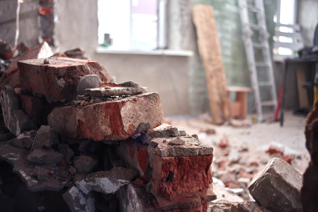 Broken bricks and rubble are piled on the floor of a partially demolished room, with tools, a ladder, and construction materials visible in the background near a window.