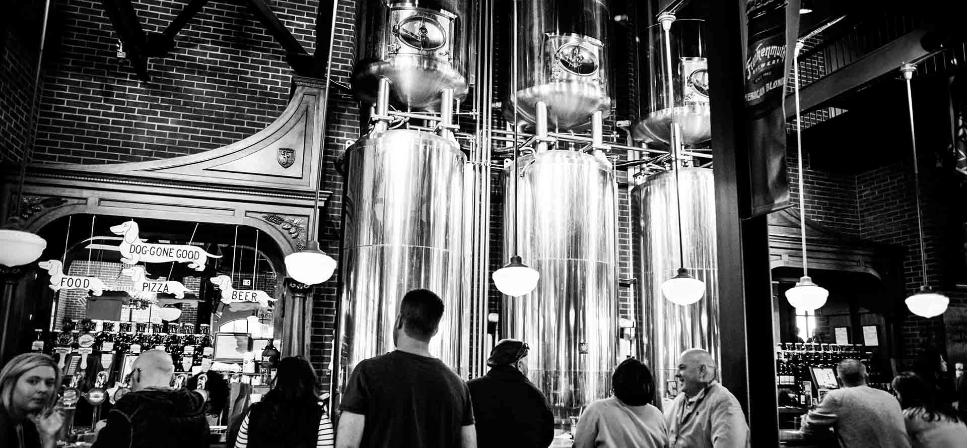 Black and white photo of people gathered inside a brewery, with tall metal brewing tanks, hanging lights, and a bar area in the background. Patrons stand and chat, and a sign advertises pizza and beer.
