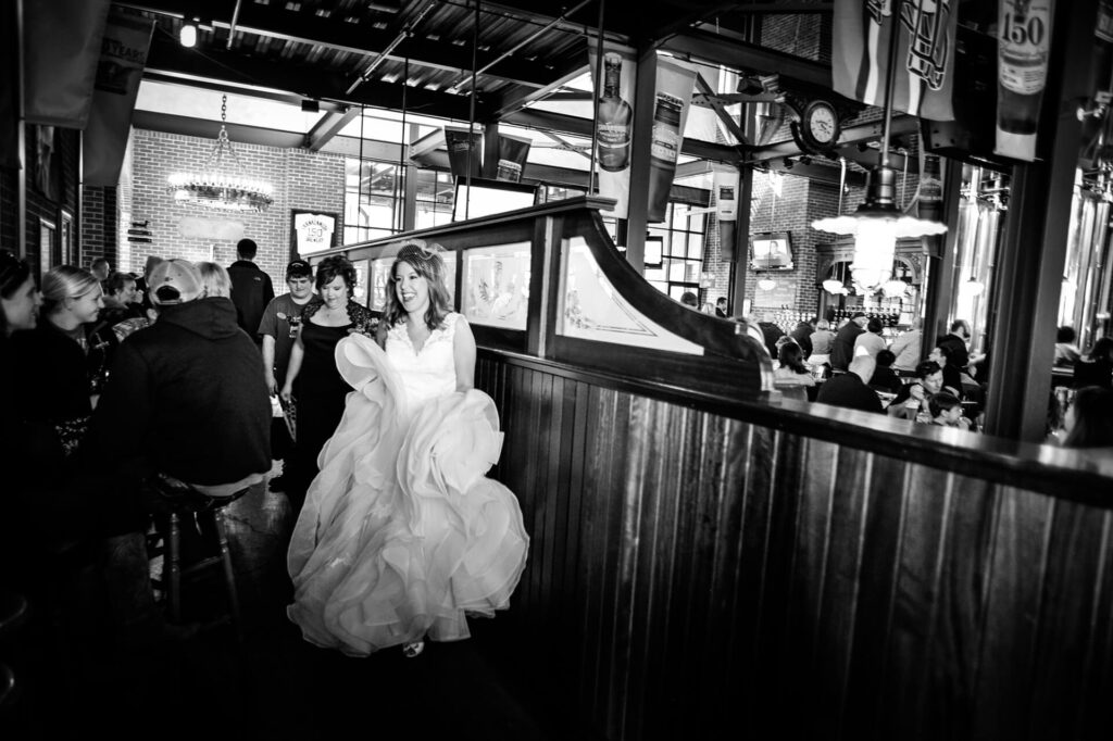 A bride in a voluminous wedding dress walks through a busy, lively restaurant or bar, smiling, with guests seated around her. The setting is industrial with brick walls, wood paneling, and hanging signs.