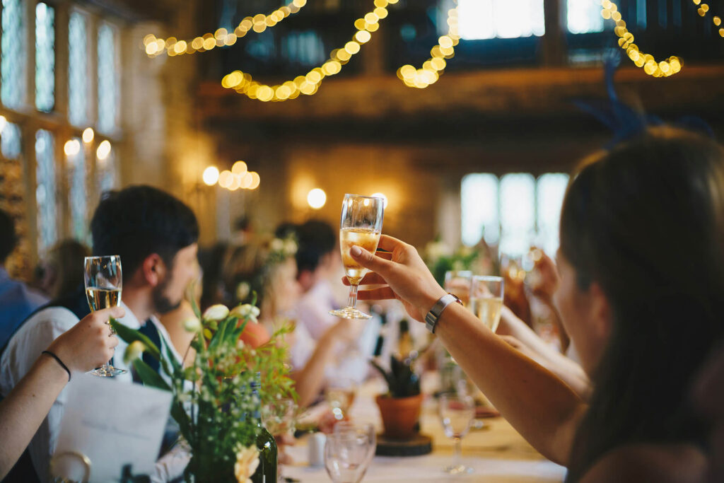 People raise glasses for a toast at a festive indoor gathering with warm lighting, floral centerpieces, and string lights creating a celebratory atmosphere.
