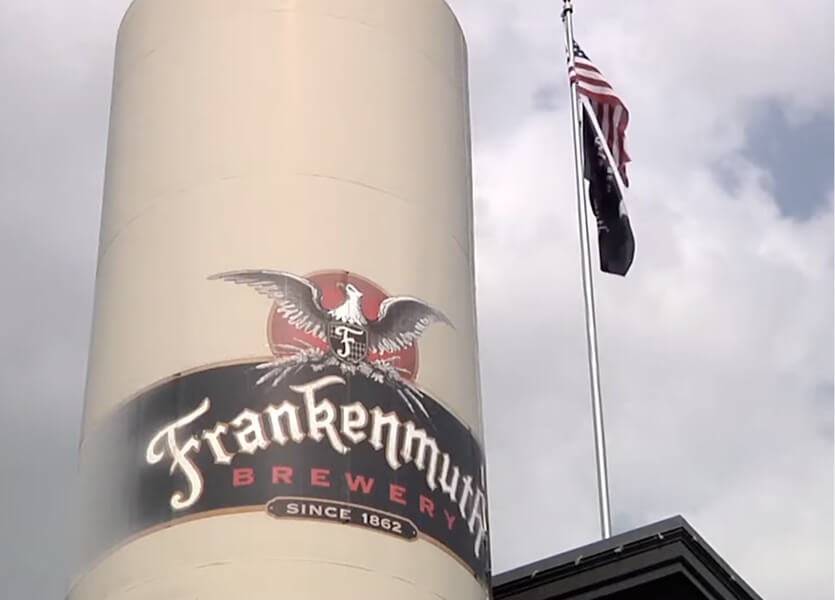 A tall tank with the Frankenmuth Brewery logo, featuring an eagle, stands under a cloudy sky beside a flagpole flying the American flag.